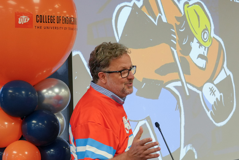 Individual speaks at a podium during a UTEP Engineering homecoming gathering while wearing a themed jersey-style shirt, with a projected UTEP Engineering graphic and balloon decorations visible beside the presentation area.
